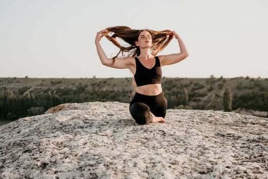 Well looking middle aged woman with long hair, fitness instructor in leggings and tops doing stretching and pilates on the rock near forest. Female fitness yoga routine concept. Healthy lifestyle.