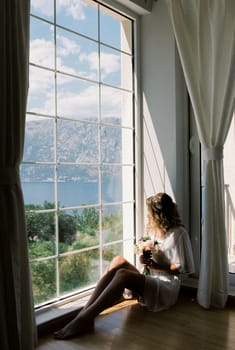 Bride in a dressing gown with a bouquet sits on the floor and looks out the window. High quality photo