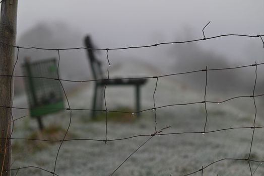 Park bench and a dustbin behind a sheep fence on a dike on a foggy morning in Wintertime