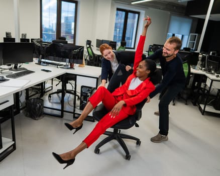 Caucasian red-haired woman, bearded caucasian man rolled African American young woman on office chair. Colleagues have fun at work