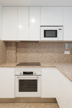 Modern kitchen is well lit by daylight from a sliding sash window. Beige granite walls with black spots. Kitchen furniture made of wood covered with white glossy paint. Built-in modern technology