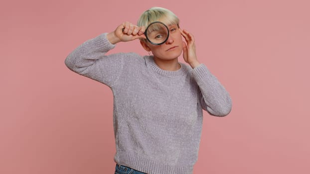 Investigator researcher scientist woman holding magnifying glass near face, looking into camera with big zoomed funny eyes, searching, analysing. Young girl on pink background. Lgbt gay lesbian people
