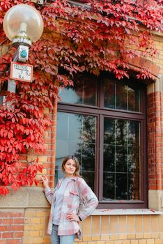 a beautiful girl stands against the background of the window of an old European house, entwined with a floating red color in autumn. Tourism & Travel Concept. Nice portrait of a young woman, in boho style outdoors in fall autumn day. Caucasian female girl 20 30 years old