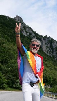 gray-haired senior elderly Caucasian man bisexuality, diabetic with a beard and sunglasses with a rainbow LGBTQIA peace flag in mountains. Celebrates Pride Month, Rainbow Flag Day, gay parade. Vertical