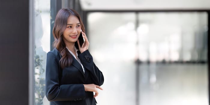 Happy young asian business woman wearing suit holding mobile phone standing in her workstation office.