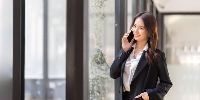 Happy young asian business woman wearing suit holding mobile phone standing in her workstation office.