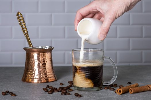 A woman's hand pours cream into a glass cup with coffee next to a traditional Turkish coffee pot. Selective focus.