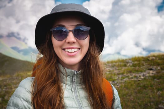 Cheerful tourist in sunglasses stands. Portrait of a traveler in the mountains. The concept of adventure, travel and hiking. Happy woman in a cap enjoys the sunshine while hiking in the mountains. Tourist with glasses.