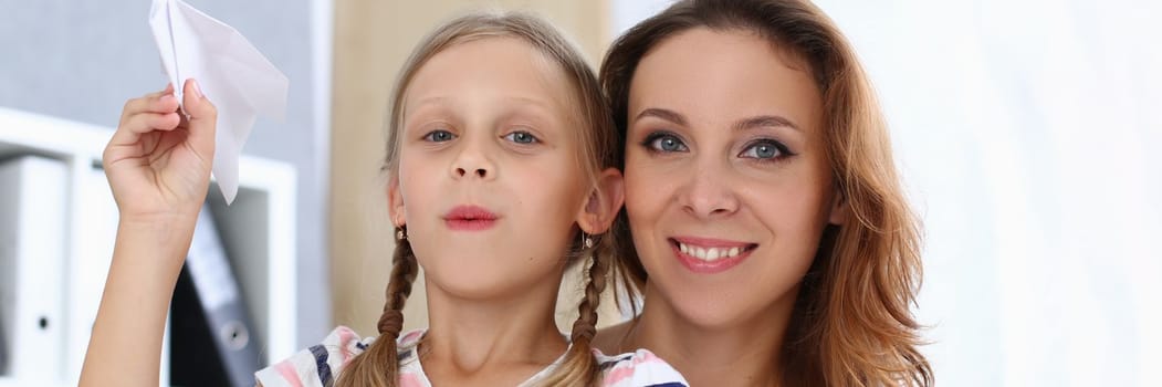 Portrait of beautiful happy mom with daughter are holding paper airplane. Home family pastime and entertainment