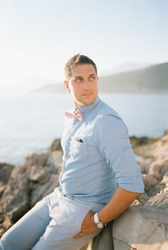 Smiling man sits on a rocky shore by the sea holding his hands in his pockets. High quality photo