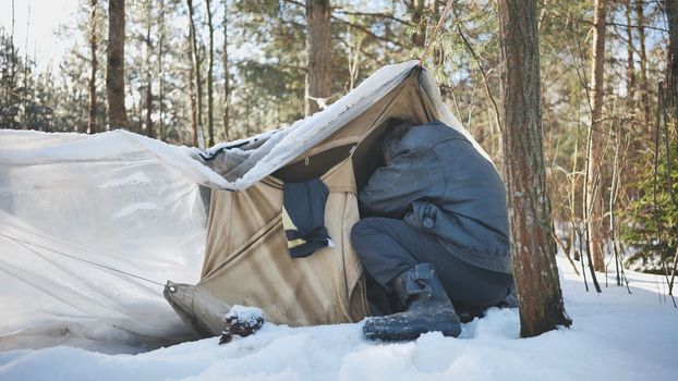 A homeless man climbs in and out of a tent in the woods in winter