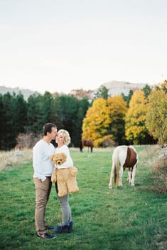 Man kisses woman with a toy bear in her hands while standing on a green lawn. High quality photo