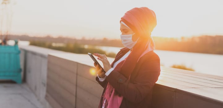 Muslim woman in hijab studying online on laptop using internet outdoor. Arab female wears medical mask while illness