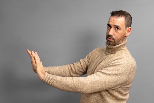 Side view of serious man making stop gesture showing palm, conflict ban warning about danger, stop bullying, wears beige turtleneck sweater. Indoor studio shot isolated on gray background.