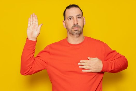 I swear to be honest. Sincere responsible handsome bearded hispanic man raising hand to take oath, promising to be honest and tell the truth, keeping hand on chest isolated on yellow background