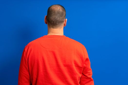 Man in his 40s with a red sweatshirt and very short hair posing with his back isolated over blue background