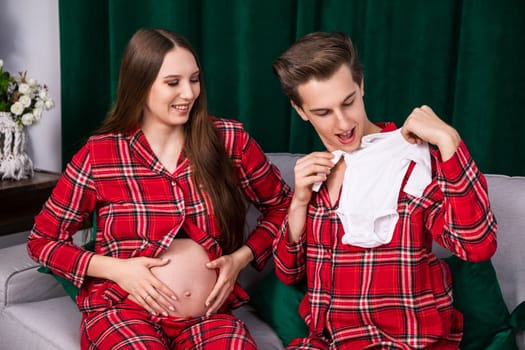 A woman stroking her pregnant belly smiles as she looks at her husband. The man sits on the couch next to his wife and displays bright baby clothes.