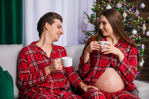 A man holds his hand on the leg of his pregnant partner. The couple is holding white teacups. In the background is a fuzzy Christmas tree and a white curtain and a green curtain.