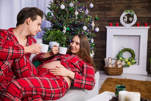 A man sitting on a couch holds the legs of a woman lying next to him on his lap. The woman is advanced in pregnancy and has an exposed belly. Both are holding white cups.