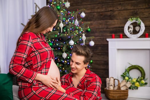 A couple on a gray couch against a background of a Christmas tree and other holiday decorations. The woman is advanced pregnant and sitting, while the man kneels in front of her placing his hand on her pregnant belly.