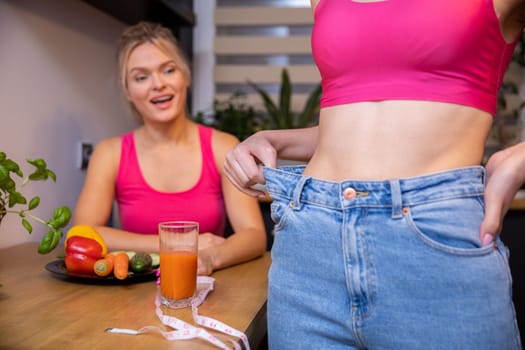 A blurry figure of a woman sitting in the background at a table looks at a friend standing in front. In the foreground, a close-up of a portion of the girl's torso with her slim belly exposed.