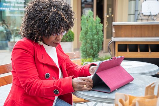 a young african american woman in a red coat sits at a table in a cafe and checking bill after shopping using a tablet, financial problems crisis, proper planning of expenses, High quality photo