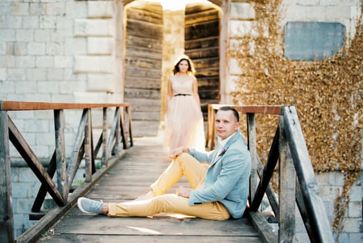 Bride walks to the groom sitting on the wooden bridge. High quality photo