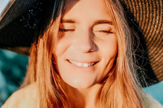 Portrait of happy young woman wearing summer black hat with large brim at beach on sunset. Closeup face of attractive girl with black straw hat. Happy young woman smiling and looking at camera at sea