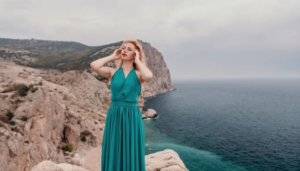 Side view a Young beautiful sensual woman in a mint long dress posing on a volcanic rock high above the sea during sunset. Girl on the nature on overcast sky background. Fashion photo