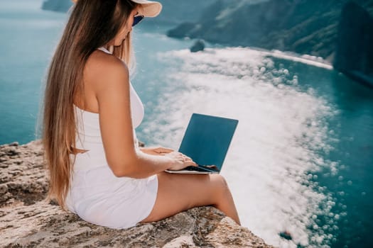 Successful business woman in yellow hat working on laptop by the sea. Pretty lady typing on computer at summer day outdoors. Freelance, travel and holidays concept.