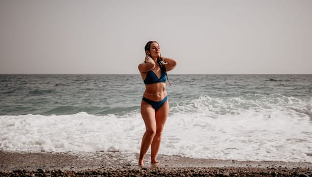Beach vacation. Hot beautiful woman in sunhat and bikini standing with her arms raised to her head enjoying looking view of beach ocean on hot summer day.