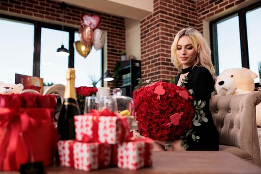 Woman holding red roses bouquet with calm facial expression in living room filled with valentines day gifts. Romantic anniversary greeting, blonde girl sitting with flowers, medium shot