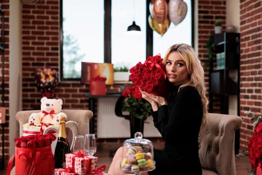 Woman holding red roses, valentines day presents. Young blonde girl with expensive flower bouquet in living room, macarons, sparkling wine, giftboxes on table, romantic love date.