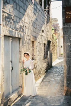Bride with a bouquet stands near the stone wall of an old house on a narrow street. High quality photo