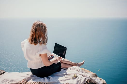 Successful business woman in yellow hat working on laptop by the sea. Pretty lady typing on computer at summer day outdoors. Freelance, travel and holidays concept.