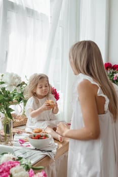 A little blonde girl with her mom on a kitchen countertop decorated with peonies. The concept of the relationship between mother and daughter. Spring atmosphere.