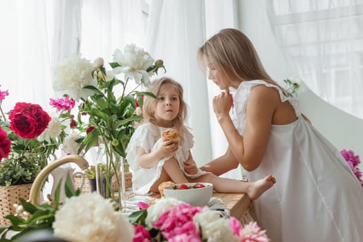 A little blonde girl with her mom on a kitchen countertop decorated with peonies. The concept of the relationship between mother and daughter. Spring atmosphere.