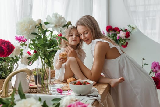 A little blonde girl with her mom on a kitchen countertop decorated with peonies. The concept of the relationship between mother and daughter. Spring atmosphere.