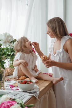 A little blonde girl with her mom on a kitchen countertop decorated with peonies. The concept of the relationship between mother and daughter. Spring atmosphere.