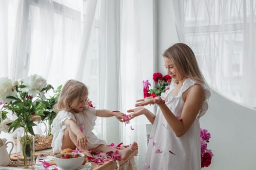 A little blonde girl with her mom on a kitchen countertop decorated with peonies. The concept of the relationship between mother and daughter. Spring atmosphere.