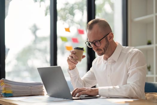 Morning coffee. young and successful business man wear eyeglasses holding cup of coffee and working with laptop while sitting at his working place.