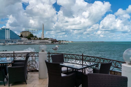 A seagull sits on an iron fence in a street cafe against the backdrop of the sea