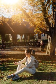 Young fashionable teenage girl with smartphone in park in autumn sitting at smiling. Trendy young woman in fall in park texting. Retouched, vibrant colors. Beautiful blonde teenage girl wearing casual modern autumn outfit sitting in park in autumn. Retouched, vibrant colors, brownish tones.