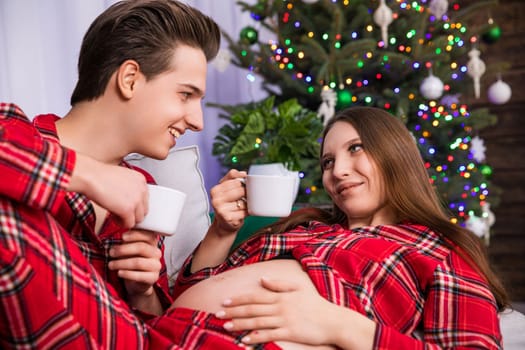 A woman in advanced pregnancy shows off her exposed pregnant belly while resting her legs on her partner's knees. The couple looks into each other's eyes and holds white cups in their hands.