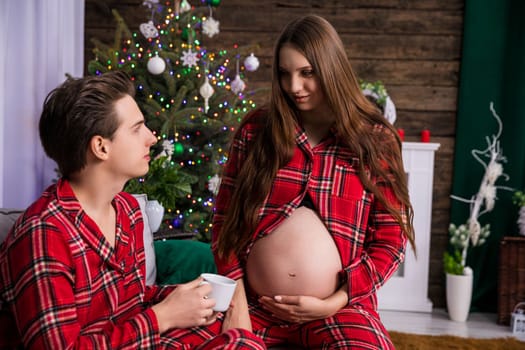 A man sits on a gray couch, while a woman with an exposed pregnant belly kneels beside him. The couple looks at each other and holds white cups in their hands. A decorated Christmas tree stands in the background.