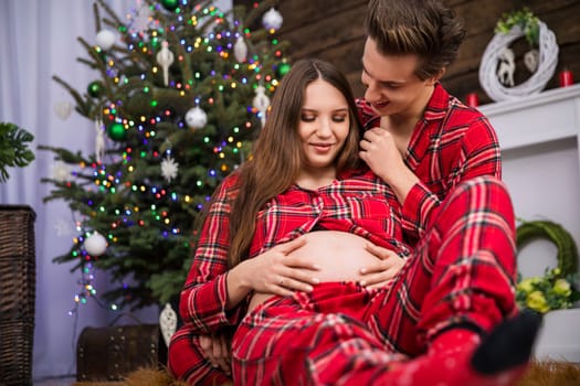 A pregnant woman sits with her legs straight leaning with her back against the man sitting behind her. They are both smiling and wearing identical red checkered pajamas.