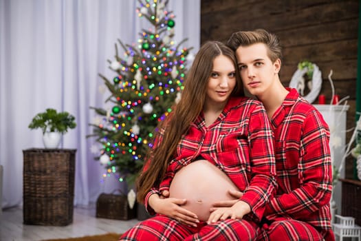 A pregnant woman and her husband sit together wearing red pajamas. A Christmas tree with lights can be seen in the background. Two people are smiling.