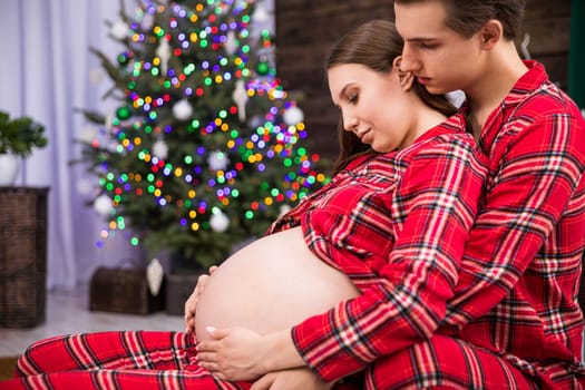 A pregnant woman leans with her back against a man sitting behind her. The man hugs the woman and holds his hand on her stomach. The hugging couple can be seen in profile against a smudged Christmas tree.