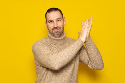 Bearded hispanic man in his 40s wearing a beige turtleneck clapping his hands in satisfaction with his hands to the side, isolated over yellow background