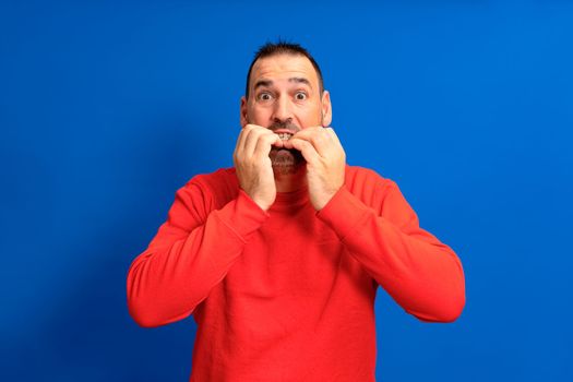 Handsome man wearing red jumper over isolated blue background looking stressed and nervous with hands on mouth biting nails. Anxiety problem.